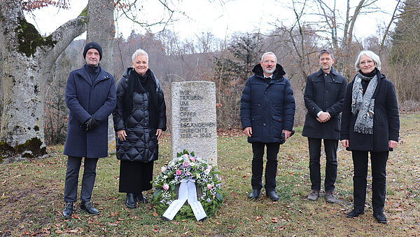 Gedenkfeier in Zwiefalten1 An einem Gedenkstein auf einer Wiese stehen rechts und links verschiedene Personen. An dem Stein liegt ein Gedenkkranz. Im Hintergrund sind Bäume.