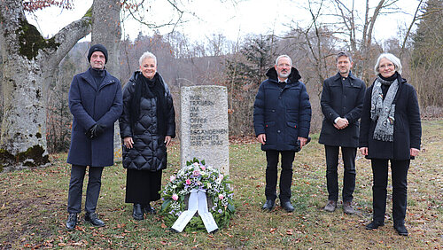 Gedenkfeier in Zwiefalten1 An einem Gedenkstein auf einer Wiese stehen rechts und links verschiedene Personen. An dem Stein liegt ein Gedenkkranz. Im Hintergrund sind Bäume.