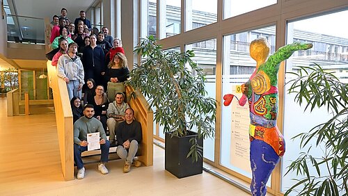Auf einer langen Treppe im Inneren eines hellen Klinikgebäudes sitzen und stehen etwa 25 Frauen und Männer in Alltagskleidung und lächeln in Richtung Kamera.