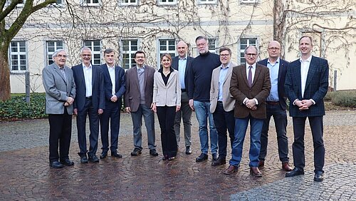 Eine Frau und zehn Männer stehen vor der Fassade eines historischen Gebäudes zum Gruppenbild zusammen und schauen freundlich in Richtung Kamera.