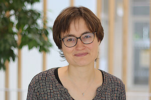 zfp-dr-sophie-hirsch.jpeg Porträtfoto einer jungen Frau. Sie hat dunkelbraune, kurze glatte Haare. Sie trägt eine runde dunkle Brille, trägt ein graues Langarmshirt und steht vor einem dunkelgrauen Hintergrund. Sie lächelt mit geöffnetem Mund in die Kamera.