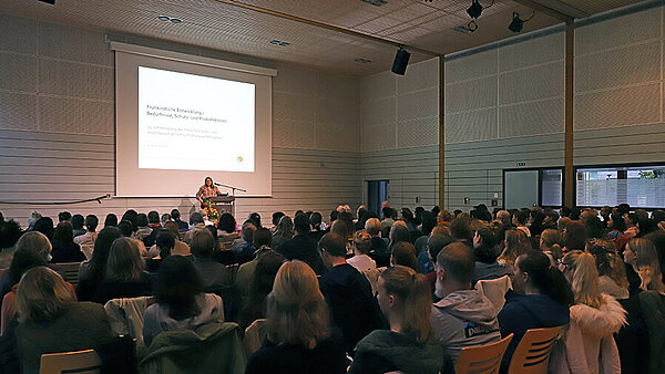 Eine vollbesetzte, bestuhlte Turnhalle. Die Zuhörer:innen blicken auf eine Frau mittleren Alters, die einen Vortrag hält. Hinter ihr an der Wand ist eine PowerPoint-Präsentation zu sehen.