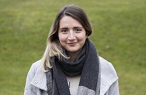 Porträtfoto einer jungen Frau mit langen braunen glatten Haaren mit Seitenscheitel. Sie trägt einen dicken dunkelgrau karierten Schal und einen hellgrauen Mantel. Sie lächelt freundlich in die Kamera. Im Hintergrund ist eine Wiese zu sehen.