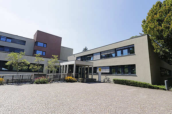 Weitwinkel-Aufnahme des Eingangsbereichs eines grauen, zwei- bzw. dreistöckigen Klinikgebäudes mit gepflastertem Vorplatz und unter blauem Himmel.