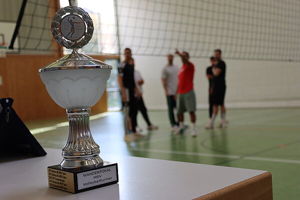 Man sieht einen großen goldenen Pokal im Vordergrund, im Hintergrund ist ein Volleyballfeld erkennbar auf dem 4 Personen beisammen stehen.