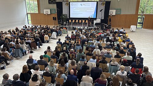 Man sieht einen Saal aus der Vogelperspektive, auf der die 350 Teilnehmenden auf Stühlen sitzen und zu einer Bühne blicken, auf der gerade ein Referent steht.
