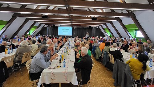 In einer vollbesetzten Halle sitzen Pensionäre an langen geschmückten Tischreihen.
