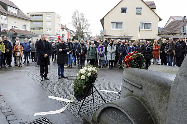 Zwei dunkel gekleidete Männer stehen in stiller Andacht vor einem Denkmal in der Form eines Omnibusses, vor ihnen stehen Gedenkkränze, um sie herum viele ebenfalls anteilnehmende Menschen.