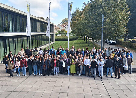 152 junge Frauen und Männer unterschiedlicher Herkunft haben sich vor einem Gebäude aufgestellt und schauen nach oben Richtung Kamera.