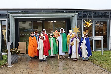 9 Kinder in bunten Gewändern und mit Kronen tragen zwei Sterne mit sich. 