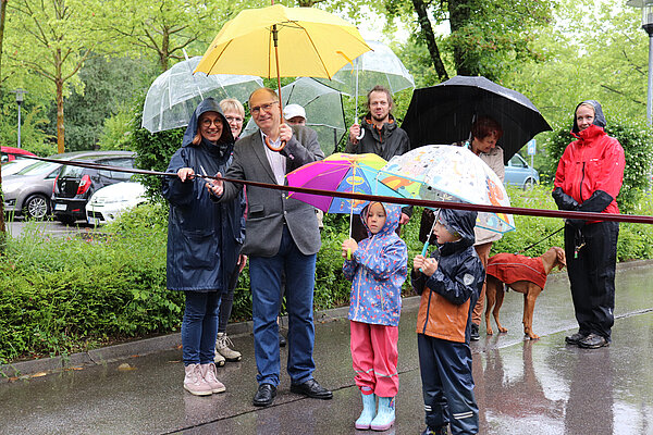 Eine Gruppe Menschen mit bunten Regenschirmen steht vor einem Stoffband. Ein Mann hält eine Schere in der Hand. 