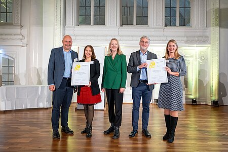 Fünf Personen stehen nebeneinander auf einer Bühne. In der Mitte befindet sich die Umweltministerin in einem grünen Sakko, rechts und links davon stehen die Unternehmensvertreter mit der Nominierungsurkunde. 