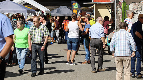 Rund 20 Menschen sind zu sehen, die bei strahlendem Sonnenschein zwischen den links und rechts befindendlichen Marktständen schlendern und sich die Produkte anschauen..