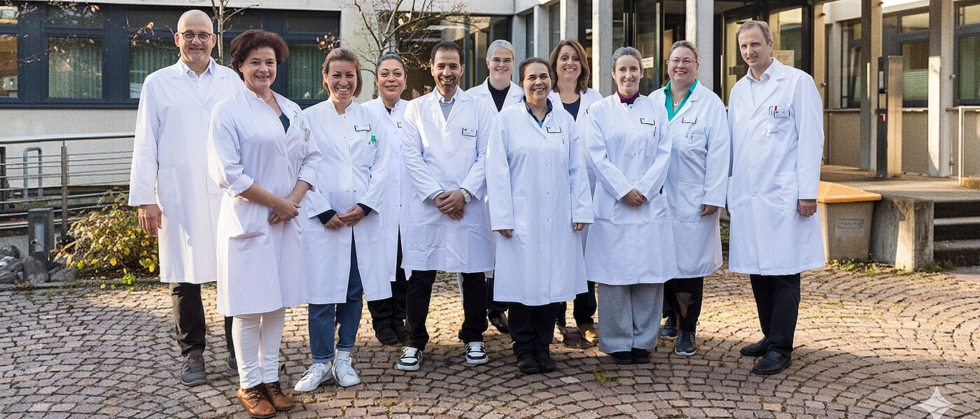 Elf Mitarbeiterinnen und Mitarbeiter der Neurologie und Epileptologie stehen freundlich lächelnd in weißen Arztkitteln vor dem Eingang zur Klinik.