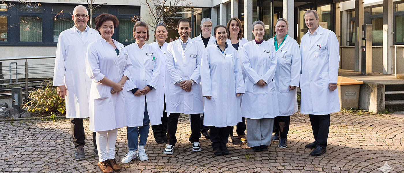Elf Mitarbeiterinnen und Mitarbeiter der Neurologie und Epileptologie stehen freundlich lächelnd in weißen Arztkitteln vor dem Eingang zur Klinik.
