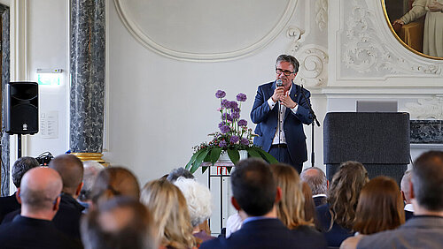 Ein Mann in blauem Anzug und weißem Hemd spricht mit Mikrofon in der Hand vor einem sitzenden Publikum in einem historischen Saal mit weißem Stuck und grauen Marmorsäulen.