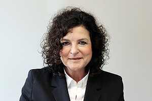 Eine Frau mit dunklen Locken, weißer Bluse und schwarzem Blazer. 