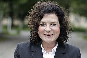 Eine Frau mit dunklen Locken, weißer Bluse und schwarzem Blazer. 