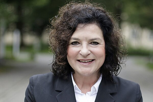 Eine Frau mit dunklen Locken, weißer Bluse und schwarzem Blazer. 