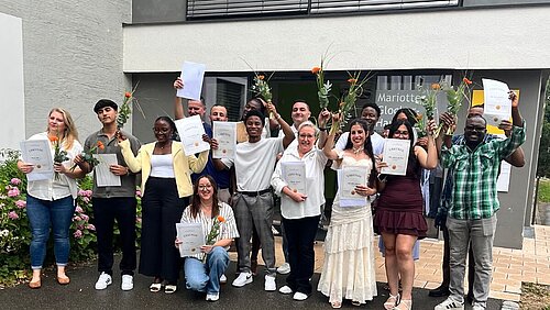 16 ausgebildete Altenpflegehelfer:innen gemischten Alters, mit und ohne Migrationshintergrund, stehen vor dem Mariotte-Glocker-Haus des ZfP Südwürttemberg und halten Zeugnisse und Blumen in den Händen.