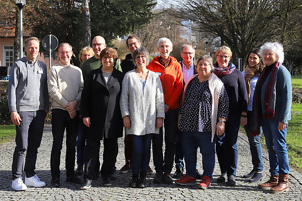 13 Frauen und Männer stehen im Freien für ein Gruppenfoto zusammen, die Sonne scheint und alle lächeln in Richtung Kamera.